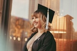A young woman wearing a graduation cap and gown is smiling while looking to the side. Her background features soft lighting with warm tones, possibly from the sun or ambient lights.