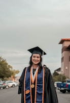 A person wearing a graduation cap and gown is standing outdoors. They are smiling and wearing a blue and orange stole with medals. In the background, there are cars parked along a street and a building is partially visible.