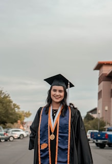 A person wearing a graduation cap and gown is standing outdoors. They are smiling and wearing a blue and orange stole with medals. In the background, there are cars parked along a street and a building is partially visible.