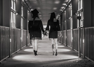 Two individuals wearing academic caps and gowns walk hand in hand down a corridor lined with windows and railings. The angle focuses on their backs, capturing a long perspective view of the hallway.
