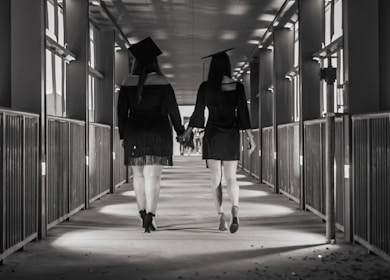 Two individuals wearing academic caps and gowns walk hand in hand down a corridor lined with windows and railings. The angle focuses on their backs, capturing a long perspective view of the hallway.