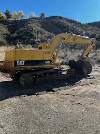 A yellow excavator is positioned in a rugged outdoor setting. The machine is sitting on a dirt surface with its arm extended, and it carries a visible 'CAT' logo. The surrounding landscape features a dry, bushy hillside, with a few trees scattered at the top and a clear blue sky.