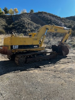 A yellow excavator is positioned in a rugged outdoor setting. The machine is sitting on a dirt surface with its arm extended, and it carries a visible 'CAT' logo. The surrounding landscape features a dry, bushy hillside, with a few trees scattered at the top and a clear blue sky.
