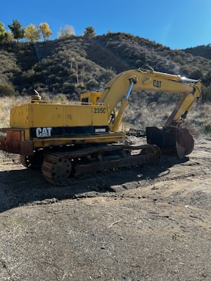 A yellow excavator is positioned in a rugged outdoor setting. The machine is sitting on a dirt surface with its arm extended, and it carries a visible 'CAT' logo. The surrounding landscape features a dry, bushy hillside, with a few trees scattered at the top and a clear blue sky.