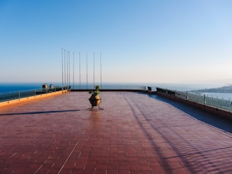 A spacious terrace with red tiles overlooks a scenic coastal view. The terrace is bordered by a decorative metal railing and features tall flagpoles and a central planter with foliage. The clear sky and distant horizon contribute to a serene atmosphere.