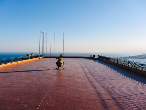 A spacious terrace with red tiles overlooks a scenic coastal view. The terrace is bordered by a decorative metal railing and features tall flagpoles and a central planter with foliage. The clear sky and distant horizon contribute to a serene atmosphere.