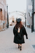 A person wearing a graduation cap and gown walks down an empty, narrow urban alleyway lined with brick buildings. The scene evokes a sense of solitude and contemplation as they move forward.