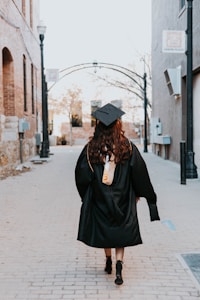 A person wearing a graduation cap and gown walks down an empty, narrow urban alleyway lined with brick buildings. The scene evokes a sense of solitude and contemplation as they move forward.