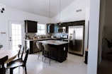 A modern kitchen featuring dark cabinetry, stainless steel appliances, and a central island with high chairs. The room is well-lit with natural light coming through a glass door and windows. A round dining table with chairs is placed on the left side. The decor is minimalistic, and the space is tidy and organized.