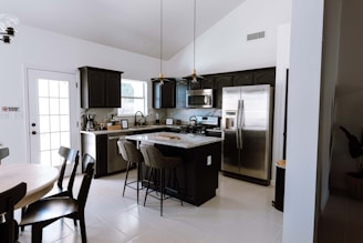 A modern kitchen featuring dark cabinetry, stainless steel appliances, and a central island with high chairs. The room is well-lit with natural light coming through a glass door and windows. A round dining table with chairs is placed on the left side. The decor is minimalistic, and the space is tidy and organized.