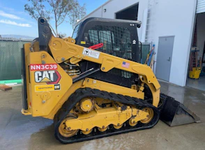 A yellow compact track loader with black tracks and a metal safety cage is parked on a wet concrete surface. The loader has a labeled CAT emblem and an American flag sticker on the side. The background shows an industrial setting with a metal fence, trees, and a building with an open door.