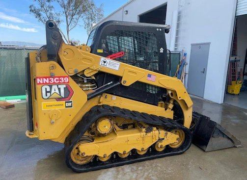 A yellow compact track loader with black tracks and a metal safety cage is parked on a wet concrete surface. The loader has a labeled CAT emblem and an American flag sticker on the side. The background shows an industrial setting with a metal fence, trees, and a building with an open door.