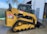 A yellow compact track loader with black tracks and a metal safety cage is parked on a wet concrete surface. The loader has a labeled CAT emblem and an American flag sticker on the side. The background shows an industrial setting with a metal fence, trees, and a building with an open door.