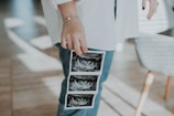 A person wearing a white shirt and blue jeans is holding several ultrasound images. The focus is on the hand and the ultrasound pictures, suggesting an intimate or meaningful context. Sunlight casts soft shadows on a wooden floor.