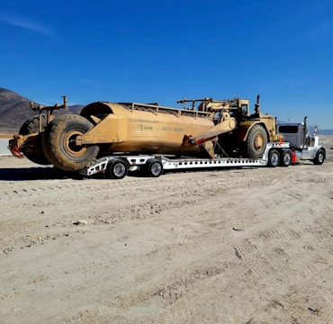 A large yellow Caterpillar water wagon is being transported by a white semi-truck on a flatbed trailer. The vehicle is situated on a dirt road with a clear blue sky overhead. In the background, there are dry, mountainous landscapes.
