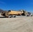A large yellow Caterpillar water wagon is being transported by a white semi-truck on a flatbed trailer. The vehicle is situated on a dirt road with a clear blue sky overhead. In the background, there are dry, mountainous landscapes.