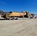 A large yellow Caterpillar water wagon is being transported by a white semi-truck on a flatbed trailer. The vehicle is situated on a dirt road with a clear blue sky overhead. In the background, there are dry, mountainous landscapes.