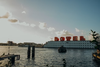 A large floating structure on the water, labeled with large red letters forming the word 'BOTEL', suggesting it is a hotel on a boat. The structure is positioned against a backdrop of clear skies with scattered clouds. The water's surface is slightly rippled, and a wooden dock extends into the water. Some industrial buildings and cranes are visible in the distance on the left side.