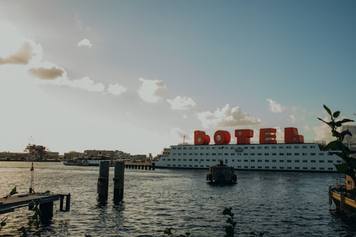 A large floating structure on the water, labeled with large red letters forming the word 'BOTEL', suggesting it is a hotel on a boat. The structure is positioned against a backdrop of clear skies with scattered clouds. The water's surface is slightly rippled, and a wooden dock extends into the water. Some industrial buildings and cranes are visible in the distance on the left side.