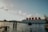 A large floating structure on the water, labeled with large red letters forming the word 'BOTEL', suggesting it is a hotel on a boat. The structure is positioned against a backdrop of clear skies with scattered clouds. The water's surface is slightly rippled, and a wooden dock extends into the water. Some industrial buildings and cranes are visible in the distance on the left side.