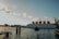 A large floating structure on the water, labeled with large red letters forming the word 'BOTEL', suggesting it is a hotel on a boat. The structure is positioned against a backdrop of clear skies with scattered clouds. The water's surface is slightly rippled, and a wooden dock extends into the water. Some industrial buildings and cranes are visible in the distance on the left side.