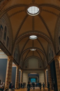 An ornate and spacious hall with an arched ceiling featuring detailed architecture and skylights, creating a grand ambiance. People are walking and standing around, suggesting a public or cultural space such as a museum or gallery.
