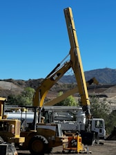 A large construction vehicle with a prominent yellow crane arm is set against a backdrop of a clear blue sky and mountainous landscape. The machinery is situated on a dirt lot, surrounded by other construction equipment including a white truck and additional vehicles. Trees and shrubs are visible along the perimeter, adding a touch of greenery to the scene.
