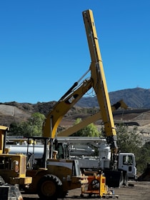 A large construction vehicle with a prominent yellow crane arm is set against a backdrop of a clear blue sky and mountainous landscape. The machinery is situated on a dirt lot, surrounded by other construction equipment including a white truck and additional vehicles. Trees and shrubs are visible along the perimeter, adding a touch of greenery to the scene.