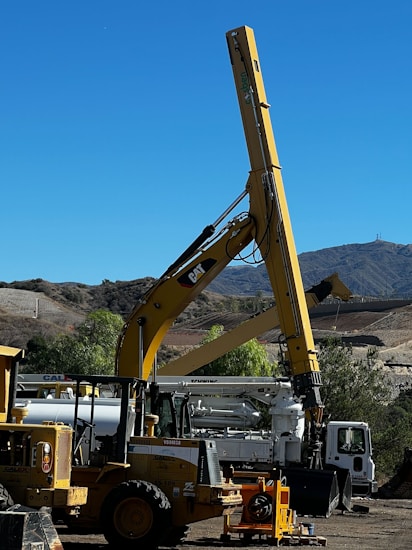 A large construction vehicle with a prominent yellow crane arm is set against a backdrop of a clear blue sky and mountainous landscape. The machinery is situated on a dirt lot, surrounded by other construction equipment including a white truck and additional vehicles. Trees and shrubs are visible along the perimeter, adding a touch of greenery to the scene.