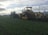 A large agricultural machine operates in a vast green field, performing tasks related to farming or land preparation. The machine features tracks and is paired with a tractor, indicating heavy-duty farm work. The sky is overcast, casting a dim light over the landscape.