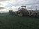 A large agricultural machine operates in a vast green field, performing tasks related to farming or land preparation. The machine features tracks and is paired with a tractor, indicating heavy-duty farm work. The sky is overcast, casting a dim light over the landscape.