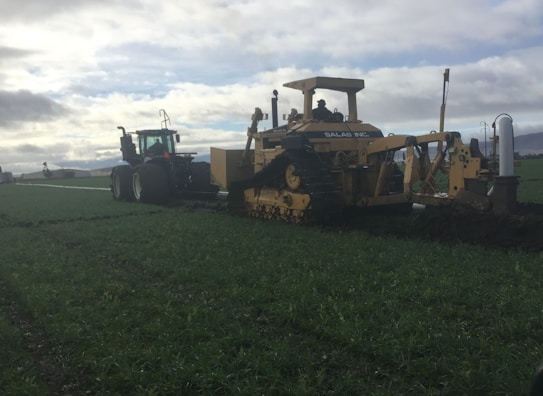 A large agricultural machine operates in a vast green field, performing tasks related to farming or land preparation. The machine features tracks and is paired with a tractor, indicating heavy-duty farm work. The sky is overcast, casting a dim light over the landscape.