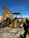 A large yellow backhoe loader equipped with a bucket on the front and an excavator arm on the back. It is parked on a gravel surface with a clear blue sky overhead. Various industrial items and fencing are visible in the background.
