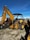 A large yellow backhoe loader equipped with a bucket on the front and an excavator arm on the back. It is parked on a gravel surface with a clear blue sky overhead. Various industrial items and fencing are visible in the background.
