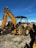 A large yellow backhoe loader equipped with a bucket on the front and an excavator arm on the back. It is parked on a gravel surface with a clear blue sky overhead. Various industrial items and fencing are visible in the background.
