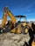 A large yellow backhoe loader equipped with a bucket on the front and an excavator arm on the back. It is parked on a gravel surface with a clear blue sky overhead. Various industrial items and fencing are visible in the background.