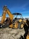 A large yellow backhoe loader equipped with a bucket on the front and an excavator arm on the back. It is parked on a gravel surface with a clear blue sky overhead. Various industrial items and fencing are visible in the background.