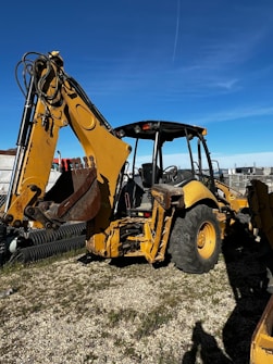 A large yellow backhoe loader equipped with a bucket on the front and an excavator arm on the back. It is parked on a gravel surface with a clear blue sky overhead. Various industrial items and fencing are visible in the background.