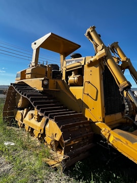 A large, yellow bulldozer with a canopy is parked on a grassy area. The machinery is detailed with metal treads and hydraulic components. A clear blue sky forms the background, while power lines run across the horizon.