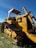 A large, yellow bulldozer with a canopy is parked on a grassy area. The machinery is detailed with metal treads and hydraulic components. A clear blue sky forms the background, while power lines run across the horizon.