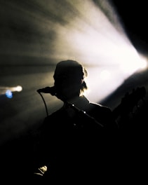 Silhouette of a person with a microphone against a backdrop of bright stage lights. The light beams radiate outward, creating a dramatic effect, while the figure remains in shadow, suggesting a musical performance.