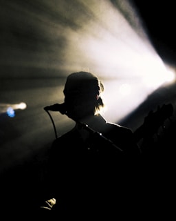 Silhouette of a person with a microphone against a backdrop of bright stage lights. The light beams radiate outward, creating a dramatic effect, while the figure remains in shadow, suggesting a musical performance.