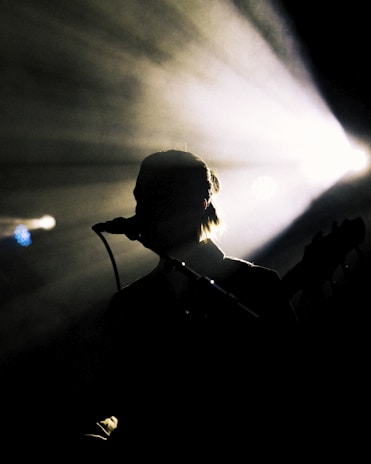 Silhouette of a person with a microphone against a backdrop of bright stage lights. The light beams radiate outward, creating a dramatic effect, while the figure remains in shadow, suggesting a musical performance.