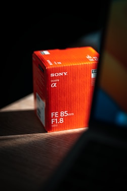 A red box for a Sony camera lens sits on a wooden surface, partially illuminated by light. The box features product specifications including 'FE 85mm F1.8' and has the Sony alpha logo prominently displayed. A blurred background and shadow create a dramatic lighting effect.