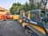 A group of construction vehicles, including skid steers and compact track loaders, are parked on a paved area. The machinery is branded with names such as Deere and Kubota. In the background, there are lush green trees and a building with a peach-colored exterior.