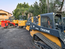 A group of construction vehicles, including skid steers and compact track loaders, are parked on a paved area. The machinery is branded with names such as Deere and Kubota. In the background, there are lush green trees and a building with a peach-colored exterior.