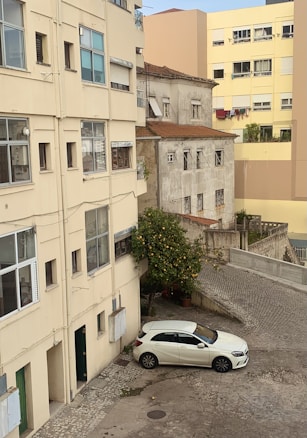 A residential area with multiple buildings, featuring peeling paint and worn exteriors. A small courtyard includes a white car parked on a cobblestone surface. A lush lemon tree grows in a corner, contrasting with the urban environment.