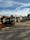 An outdoor industrial worksite is shown with a forklift in the foreground. The area contains piles of wooden planks and other materials. In the background, there are structures including storage containers and various equipment.