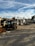An outdoor industrial worksite is shown with a forklift in the foreground. The area contains piles of wooden planks and other materials. In the background, there are structures including storage containers and various equipment.