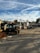 An outdoor industrial worksite is shown with a forklift in the foreground. The area contains piles of wooden planks and other materials. In the background, there are structures including storage containers and various equipment.